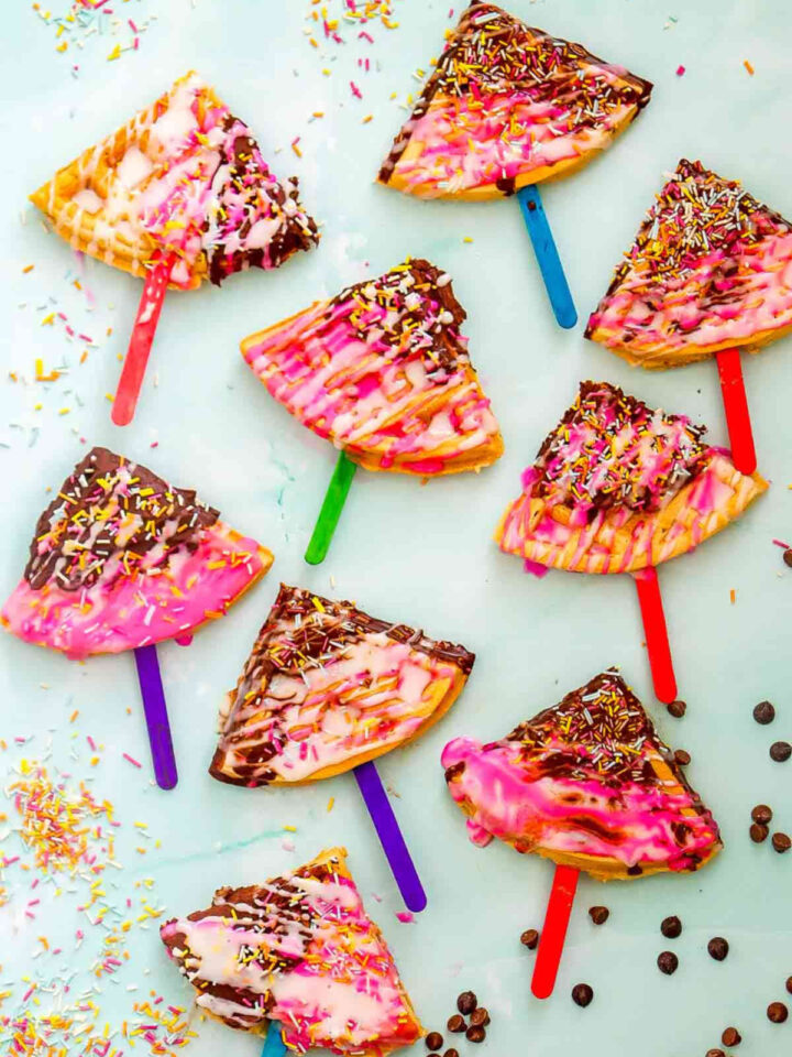 Colorful waffle pops with pink and white icing, chocolate drizzle, and rainbow sprinkles on pastel sticks.