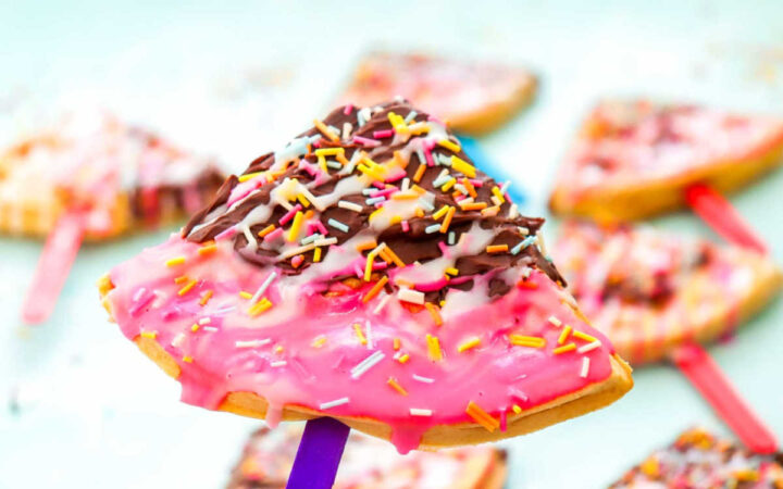 Single waffle pop with layers of pink icing, chocolate drizzle, and colorful sprinkles held by a blue stick.