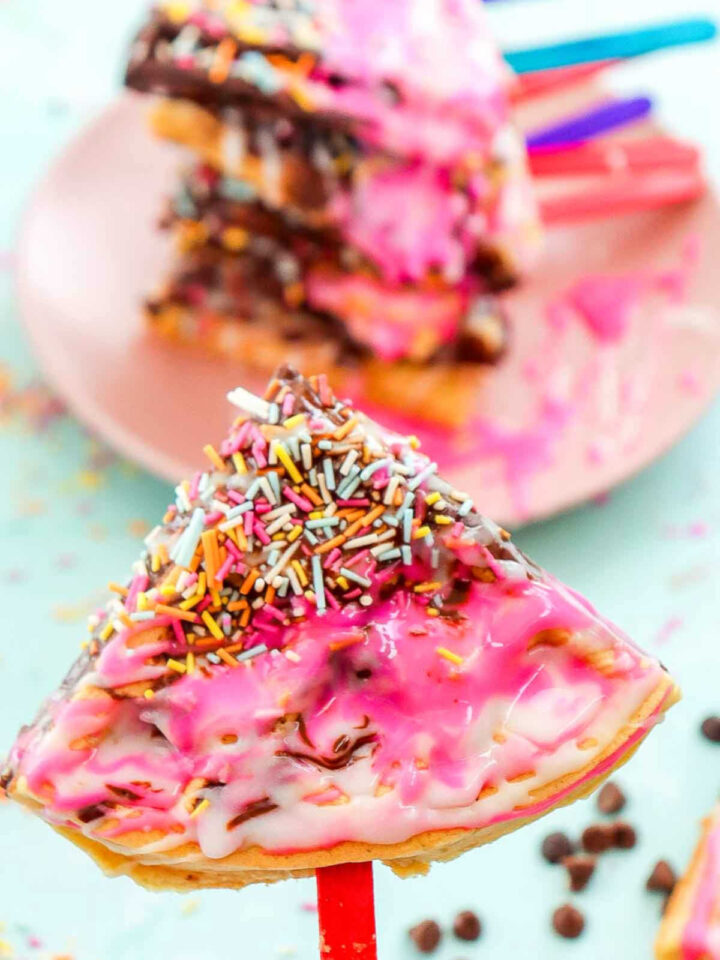 Bright waffle pop with pink icing, white drizzle, and sprinkles on a red stick with more waffle pops in the background.