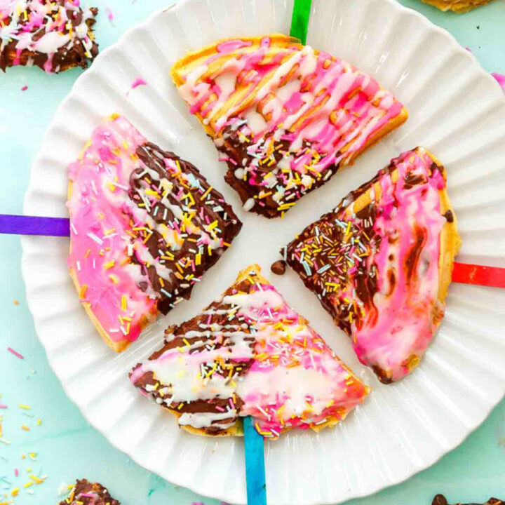 Four waffle pops on a white plate decorated with pink icing, chocolate drizzle, and rainbow sprinkles for a colorful dessert idea.