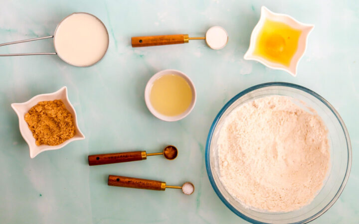 Waffle pop ingredients on a light blue background with flour, milk, egg, sugar, oil, and measuring spoons arranged neatly.