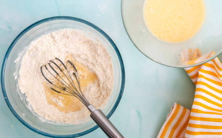 Mixing flour and egg mixture in a glass bowl with a whisk to prepare waffle batter.