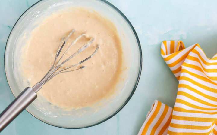Smooth waffle batter being whisked in a bowl with a yellow striped napkin beside it.