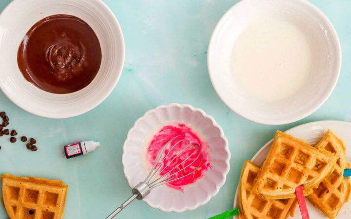 Chocolate and pink icing prepared in bowls with golden waffles and colorful sticks ready for decorating.