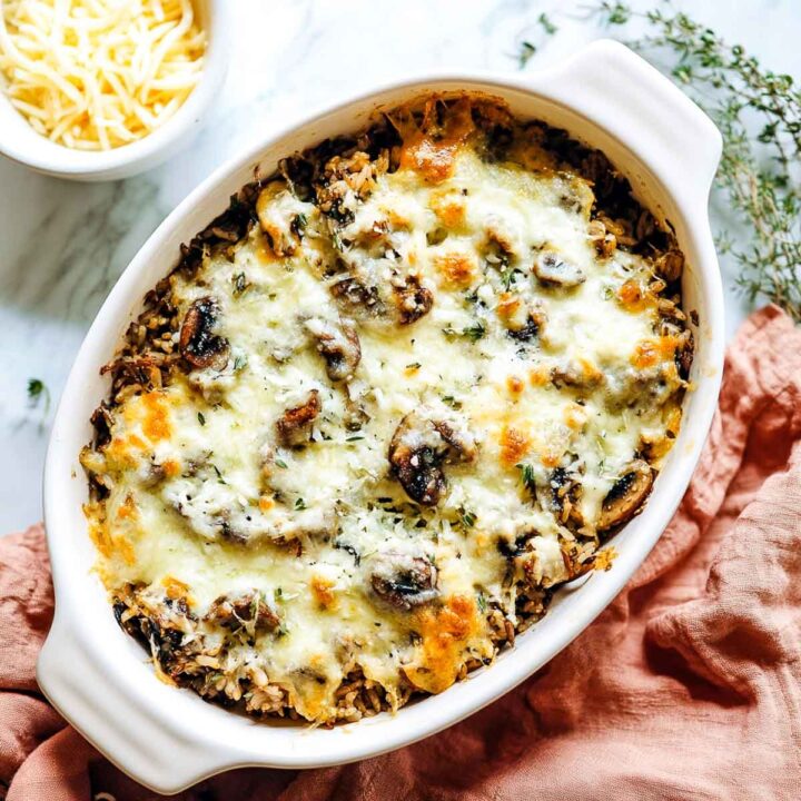 A baked wild rice and mushroom casserole in a white oval dish, topped with melted, bubbly Gruyère or Monterey Jack cheese and sprinkled with fresh thyme.