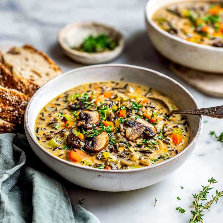 A bowl of creamy wild rice and mushroom soup, filled with sliced sautéed mushrooms, carrots, celery, and dark wild rice. It is garnished with fresh chives and parsley and served next to slices of crusty bread on a white marble surface.