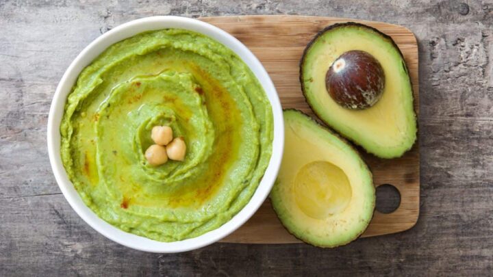 Overhead view of a bowl of bright green avocado hummus, drizzled with olive oil, garnished with whole chickpeas, and served next to a halved avocado on a wooden cutting board.