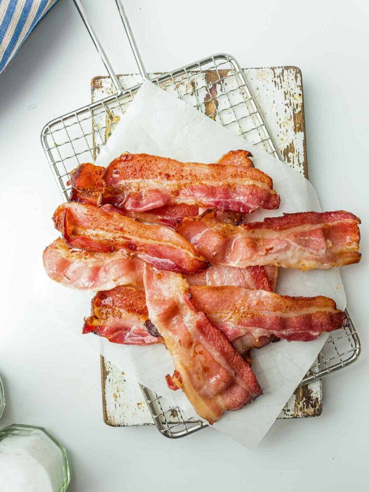 A pile of glossy, cooked bacon strips with crispy edges, resting on parchment paper on a wire rack and a distressed wooden board.