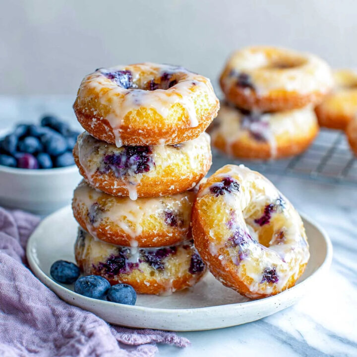 A stack of four golden-brown blueberry cake donuts topped with a white glaze, served on a plate with fresh blueberries and a purple napkin.