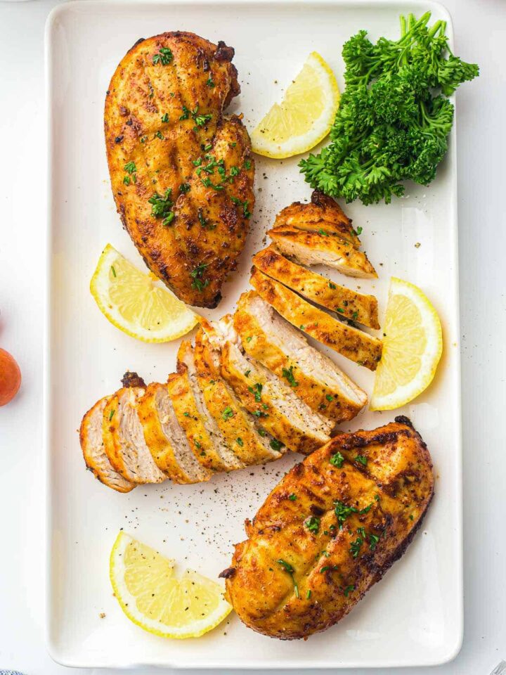 Three seasoned, cooked chicken breasts on a white rectangular platter, with one sliced into pieces, garnished with lemon wedges and fresh parsley.