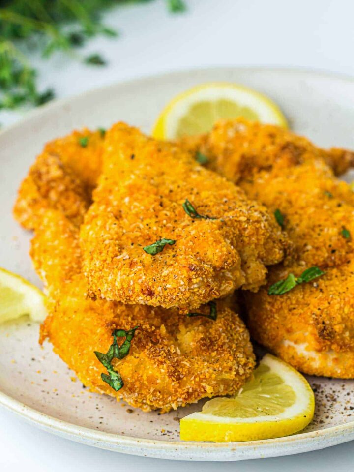 Golden-brown, breaded chicken cutlets or tenders garnished with herbs and served with fresh lemon slices on a light plate.