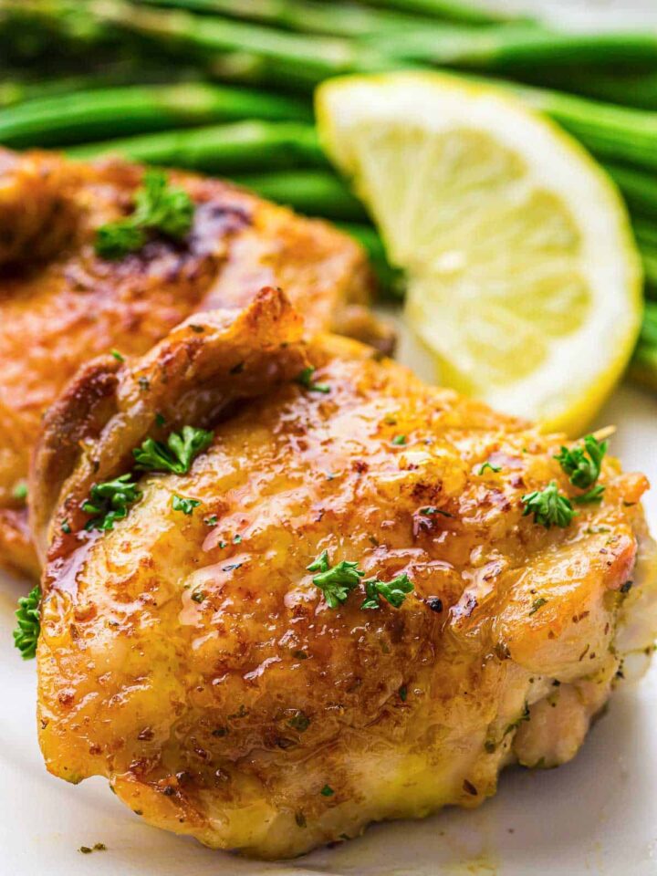 Close-up of two glazed, crispy-skinned chicken thighs garnished with parsley, served with roasted asparagus and a lemon wedge.