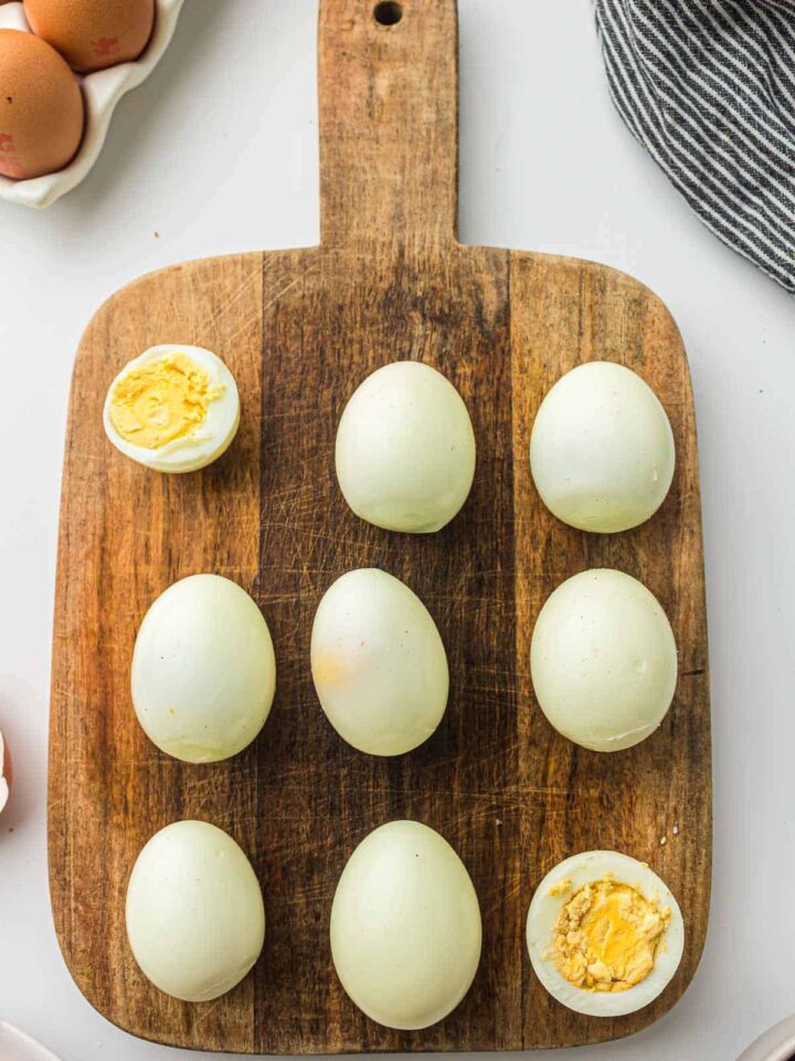 Eight peeled, whole hard-boiled eggs and two halved eggs showing firm yellow yolks, arranged on a rustic wooden cutting board.