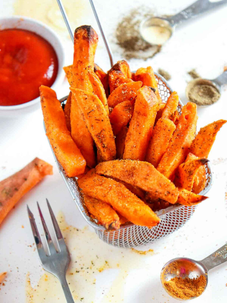 Crispy, seasoned sweet potato fries served upright in a small metal fryer basket with a bowl of ketchup and measuring spoons nearby.