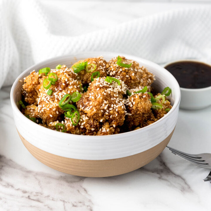 Crispy, breaded teriyaki cauliflower wings, topped with sesame seeds and green onions, served in a ceramic bowl on a marble surface.