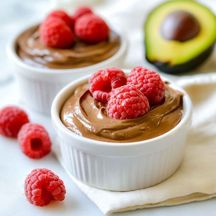 Two servings of thick avocado chocolate mousse in white ramekins, garnished with fresh raspberries.