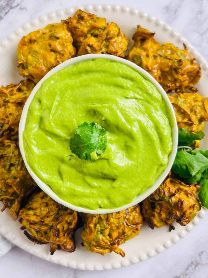 A white bowl of vibrant green avocado cilantro chutney or dip, surrounded by deep-fried pakora or vegetable fritters.