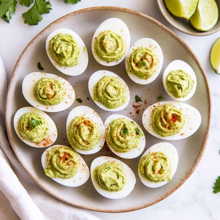 Twelve deviled eggs with a creamy green avocado yolk filling, sprinkled with paprika and cilantro, served on a round plate.