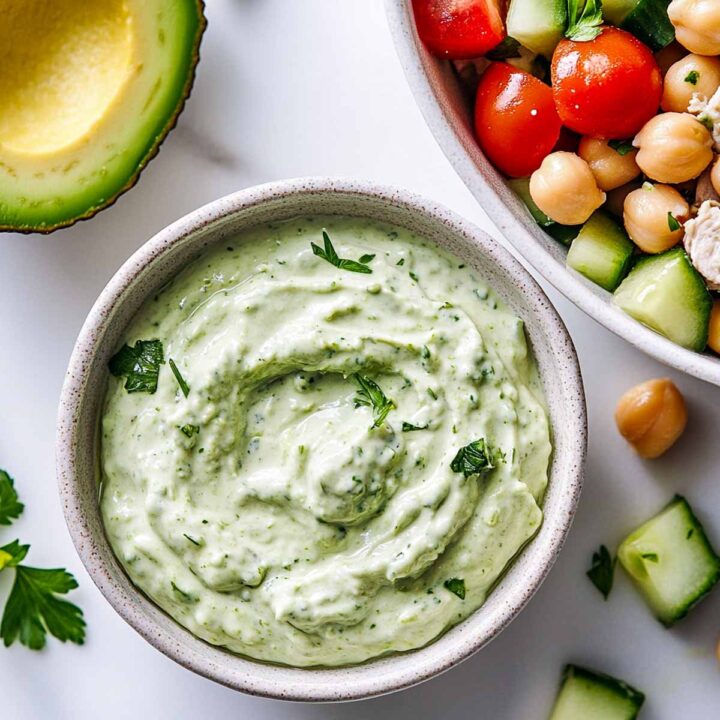 Close-up of a creamy green avocado and Greek yogurt dressing in a small bowl, with a bowl of chickpeas, tomatoes, and cucumber salad visible on the side.