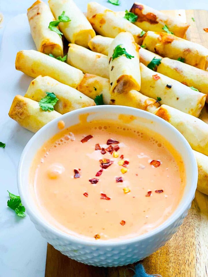 A small white bowl of creamy, orange-pink bang bang sauce, sprinkled with red pepper flakes and served with a pile of baked or fried spring rolls.