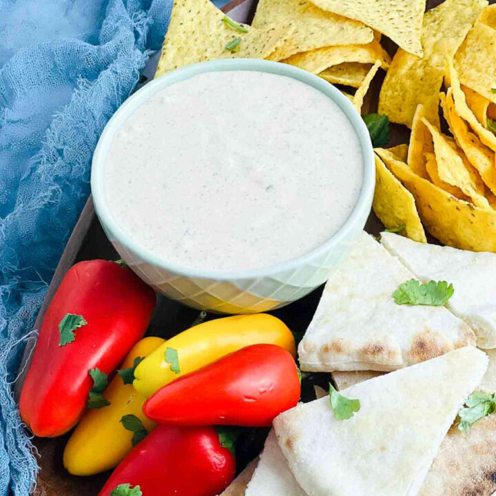 A bowl of creamy black garlic dipping sauce surrounded by tortilla chips, sliced pita bread, and whole red and yellow mini bell peppers.