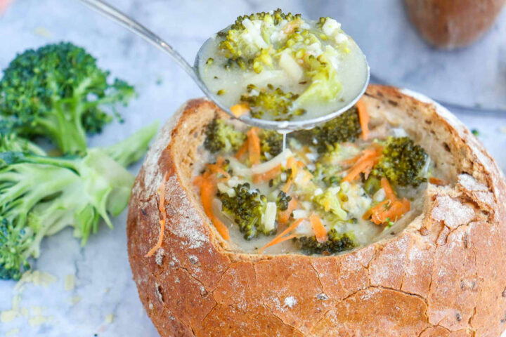 A close-up shot of a creamy broccoli cheddar soup served inside a large, crusty bread bowl, with a spoonful of the soup being lifted above the bowl, showcasing visible pieces of broccoli and shredded carrots.