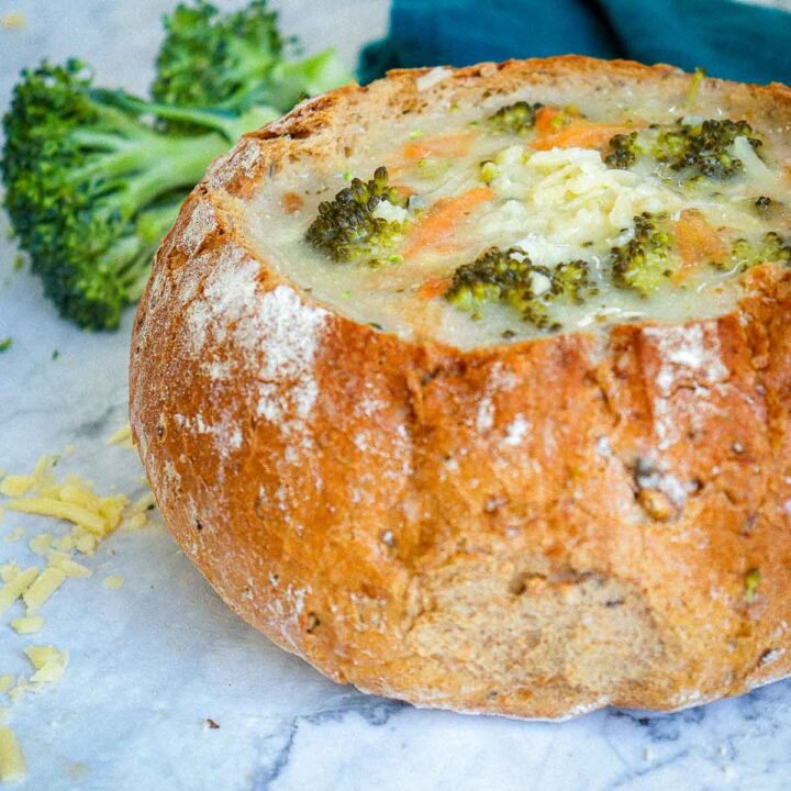A side-angle close-up of a crusty round bread bowl filled with creamy broccoli cheddar soup, topped with shredded cheese and visible broccoli florets. A large broccoli crown and shredded cheese are visible in the soft-focus background.