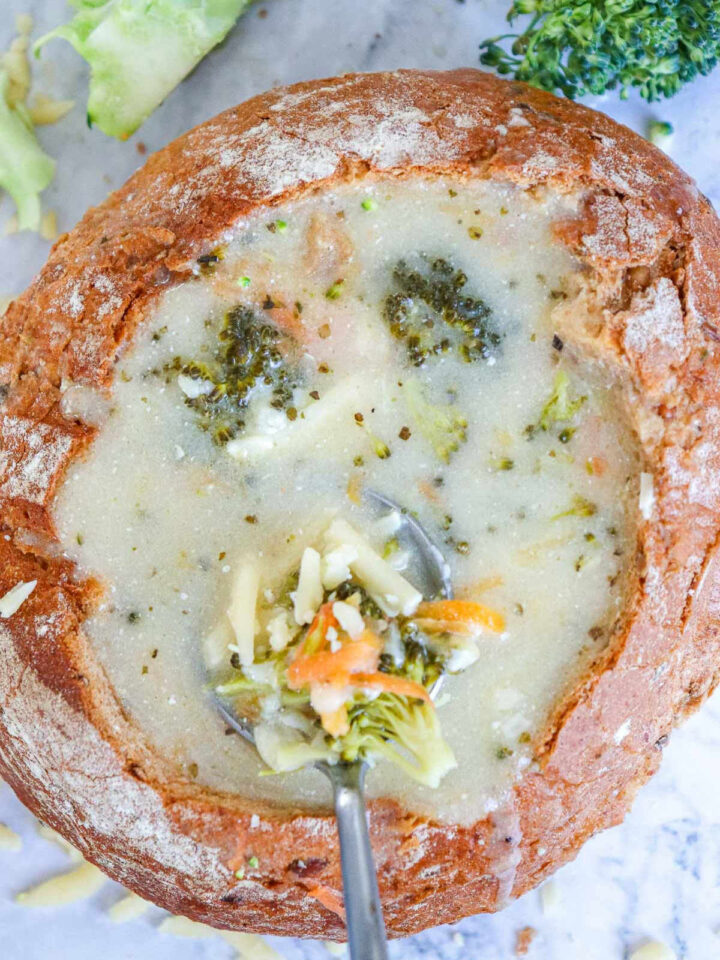 Overhead close-up of creamy broccoli cheddar soup served inside a round, crusty sourdough bread bowl, with a spoonful of soup, broccoli florets, and shredded carrots being lifted out.