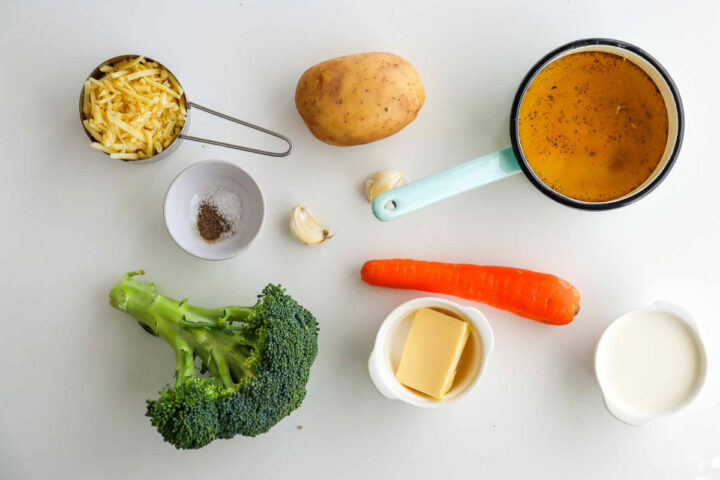 Overhead shot of ingredients for broccoli cheddar soup laid out on a white surface, including: a measuring cup of shredded cheddar cheese, one whole potato, two cloves of garlic, a light blue saucepan containing broth, one whole carrot, a small bowl with a pat of butter, a bowl of heavy cream or milk, a crown of broccoli, and a small pinch bowl of salt and pepper.