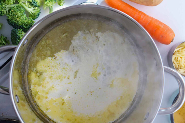 Overhead close-up of a large pot on a stovetop containing a simmering, frothy yellow liquid base for soup, likely broth mixed with melted butter and milk or cream. A carrot, broccoli florets, and shredded cheese are partially visible around the edges of the pot.