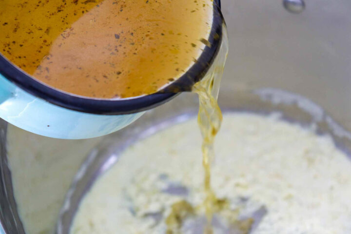 Close-up overhead shot of a light blue saucepan pouring golden-brown broth into a large pot containing a creamy, white base mixture for the soup.