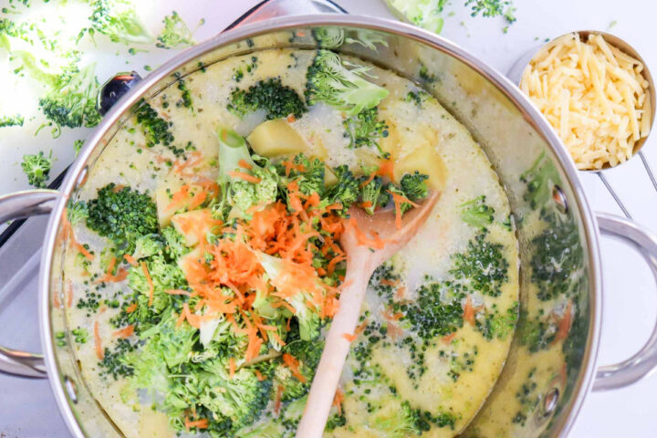 Overhead shot of a large metal pot filled with simmering cream-based soup. Fresh broccoli florets and shredded carrots have just been added to the pot and are being stirred by a wooden spoon. Shredded cheddar cheese is visible in a measuring cup on the right.