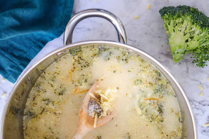 Overhead shot of a pot of finished broccoli cheddar soup simmering, with a wooden spoon holding a small amount of shredded cheese and seasoning (salt/pepper) being dipped into the liquid. A broccoli crown sits on the marble countertop to the right.