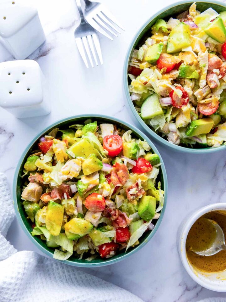 Overhead shot of two teal bowls filled with a colorful chopped salad, featuring mixed lettuce, diced chicken, bacon bits, cherry tomatoes, avocado, and red onion. A small cup of vinaigrette dressing and a set of forks are visible on the marble surface.