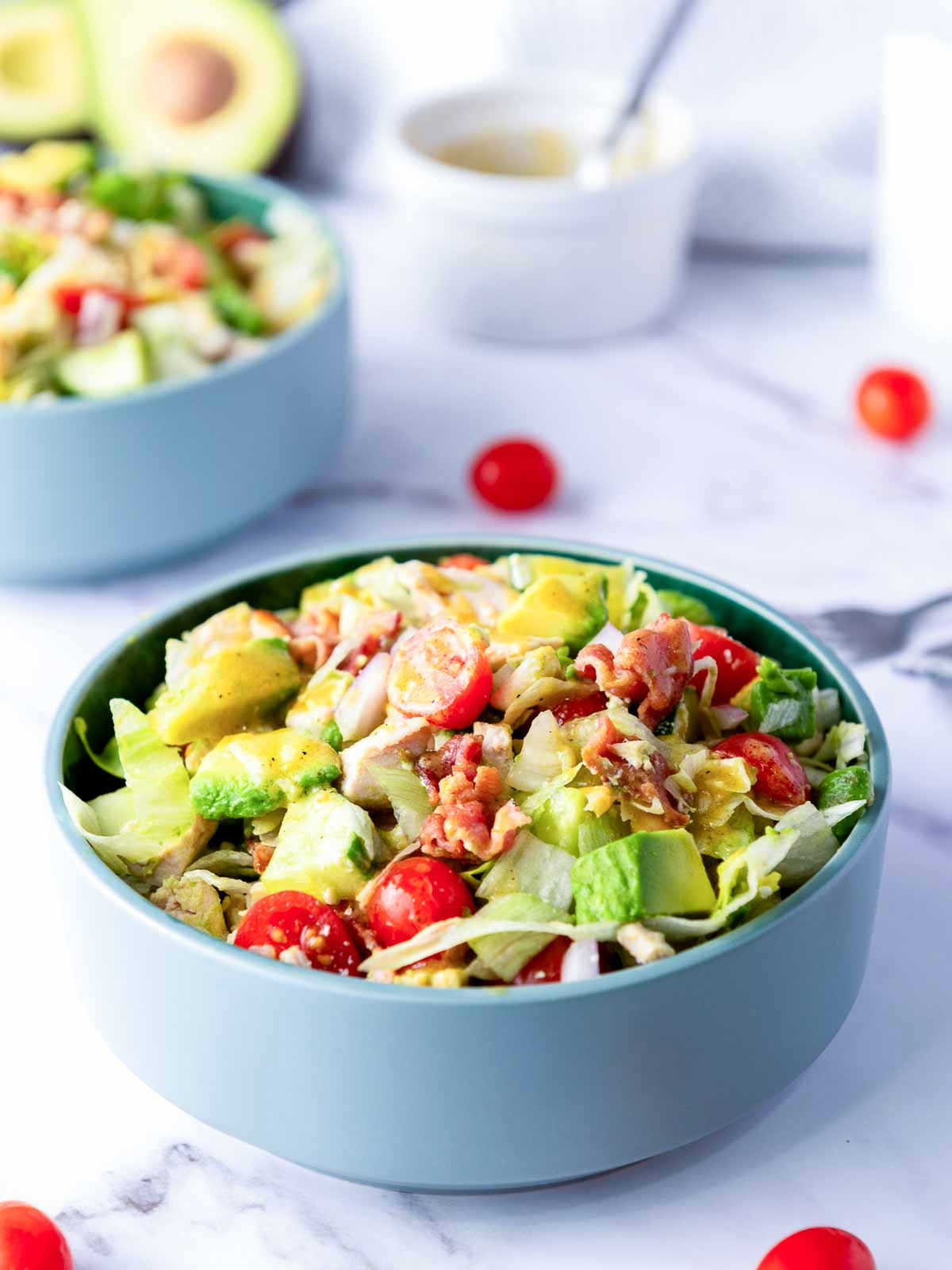 The image shows a vibrant, freshly prepared Chopped Salad served in a teal blue bowl. The salad is composed of uniformly diced ingredients, including shredded lettuce, pieces of cooked chicken, diced avocado, crumbled bacon, and halved cherry tomatoes. The salad is lightly tossed in a clear vinaigrette dressing. A bowl of dressing and a knife and fork are visible in the soft-focus background, suggesting a light, healthy lunch.
