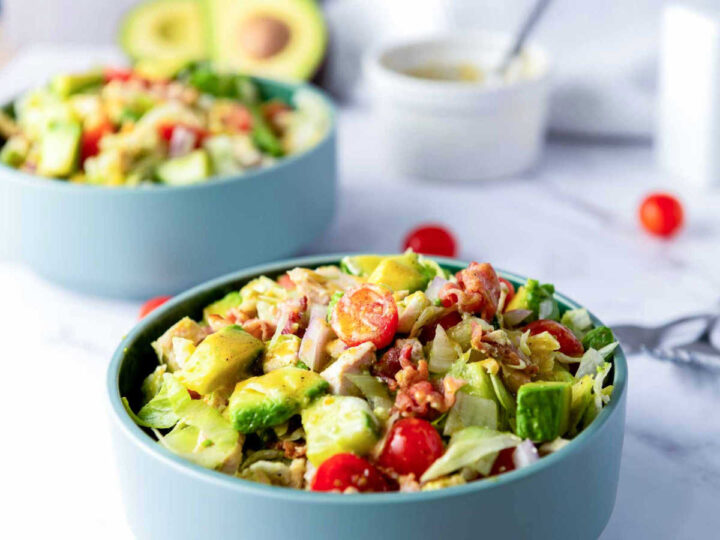 A close-up, eye-level shot of a teal bowl filled with a colorful chopped chicken salad containing lettuce, diced avocado, cherry tomatoes, and crumbled bacon, with a second bowl and a small white dish of dressing blurred in the background.