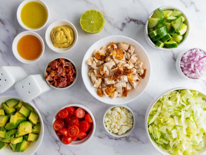 Overhead flat lay of ingredients for a chopped chicken salad laid out on a marble surface, including a central bowl of chopped cooked chicken, surrounded by bowls of diced avocado, halved cherry tomatoes, crumbled bacon, crumbled cheese (feta or blue cheese), shredded lettuce, diced cucumber, minced red onion, mustard, two small bowls of oil/vinegar, and a halved lime.