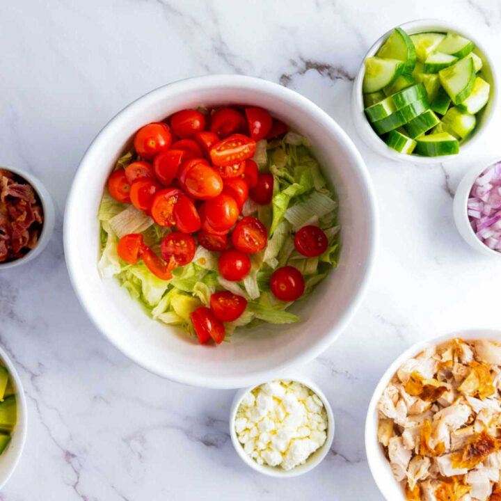 Overhead shot showing the first step of building a chopped salad: a large white bowl containing a base layer of shredded lettuce topped with halved cherry tomatoes. Surrounding the bowl are smaller bowls of diced cucumber, red onion, crumbled cheese, cooked chicken, bacon bits, and avocado.
