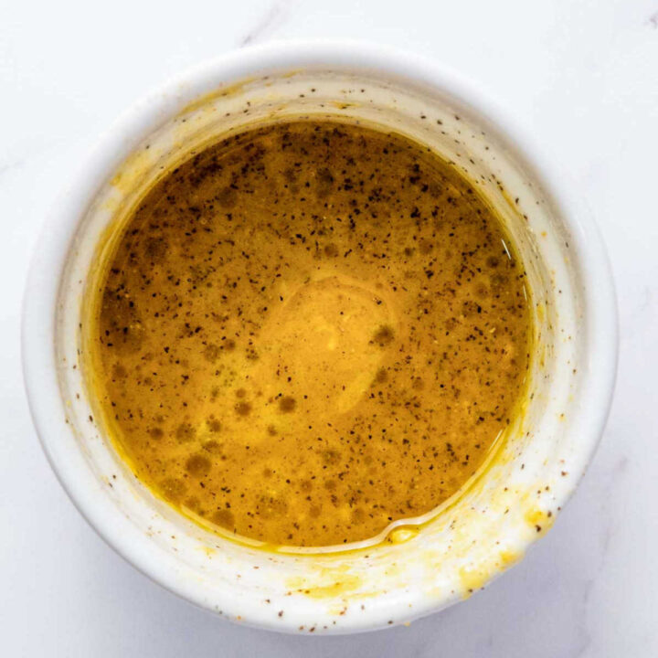 Overhead close-up of a small white bowl containing a well-mixed, emulsified vinaigrette dressing, appearing yellow-gold with specks of black pepper and possibly mustard, ready to be poured over a salad.
