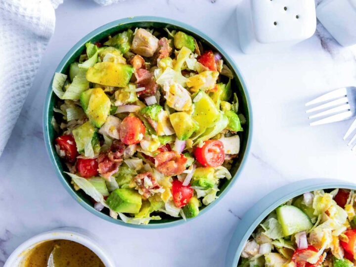 Overhead shot of a teal bowl filled with a fully tossed chopped chicken salad, showing the mixed ingredients (lettuce, chicken, avocado, bacon, tomatoes, and onion) coated in a light vinaigrette dressing. A second bowl of salad and a container of dressing are visible in the foreground.