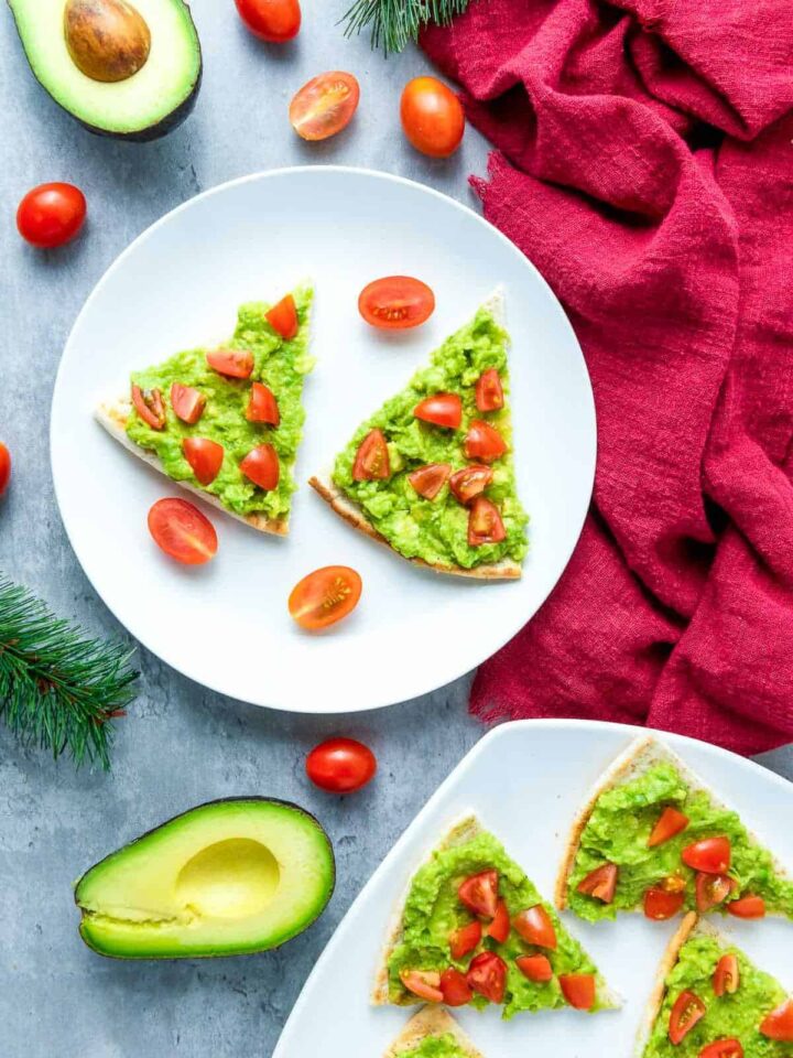 Pita bread or toast cut into a Christmas tree shape, topped with mashed avocado and chopped cherry tomatoes, and served on white plates.