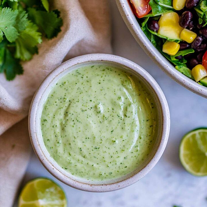 A small bowl of creamy cilantro lime Greek yogurt dressing, placed next to a southwest-style salad and lime slices.