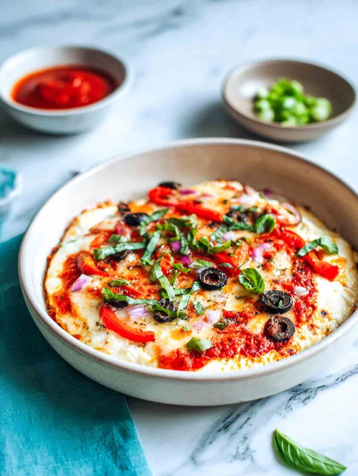 Cheesy cottage cheese pizza bowl with colorful vegetables and basil on marble surface.