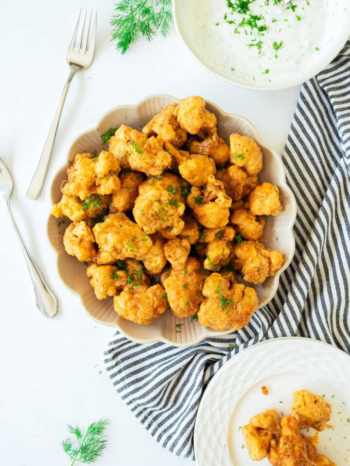 Overhead view of a generous serving of crispy, battered, and fried cauliflower bites (or wings) garnished with parsley, served in a scalloped beige bowl. A bowl of creamy white dipping sauce, forks, and a striped towel are arranged around the main dish.