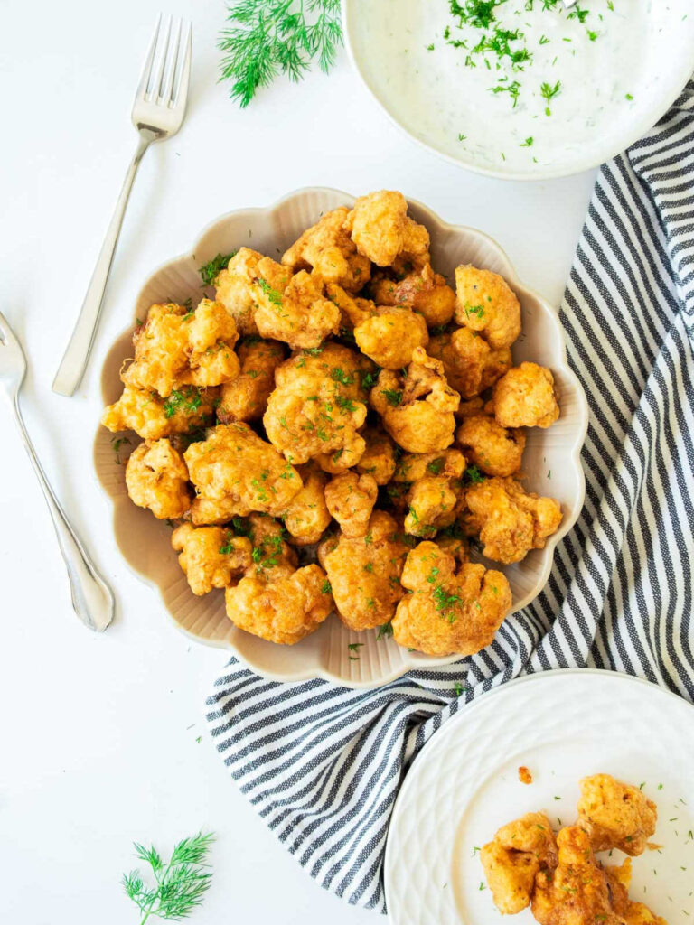 Overhead view of a generous serving of crispy, battered, and fried cauliflower bites (or wings) garnished with parsley, served in a scalloped beige bowl. A bowl of creamy white dipping sauce, forks, and a striped towel are arranged around the main dish.