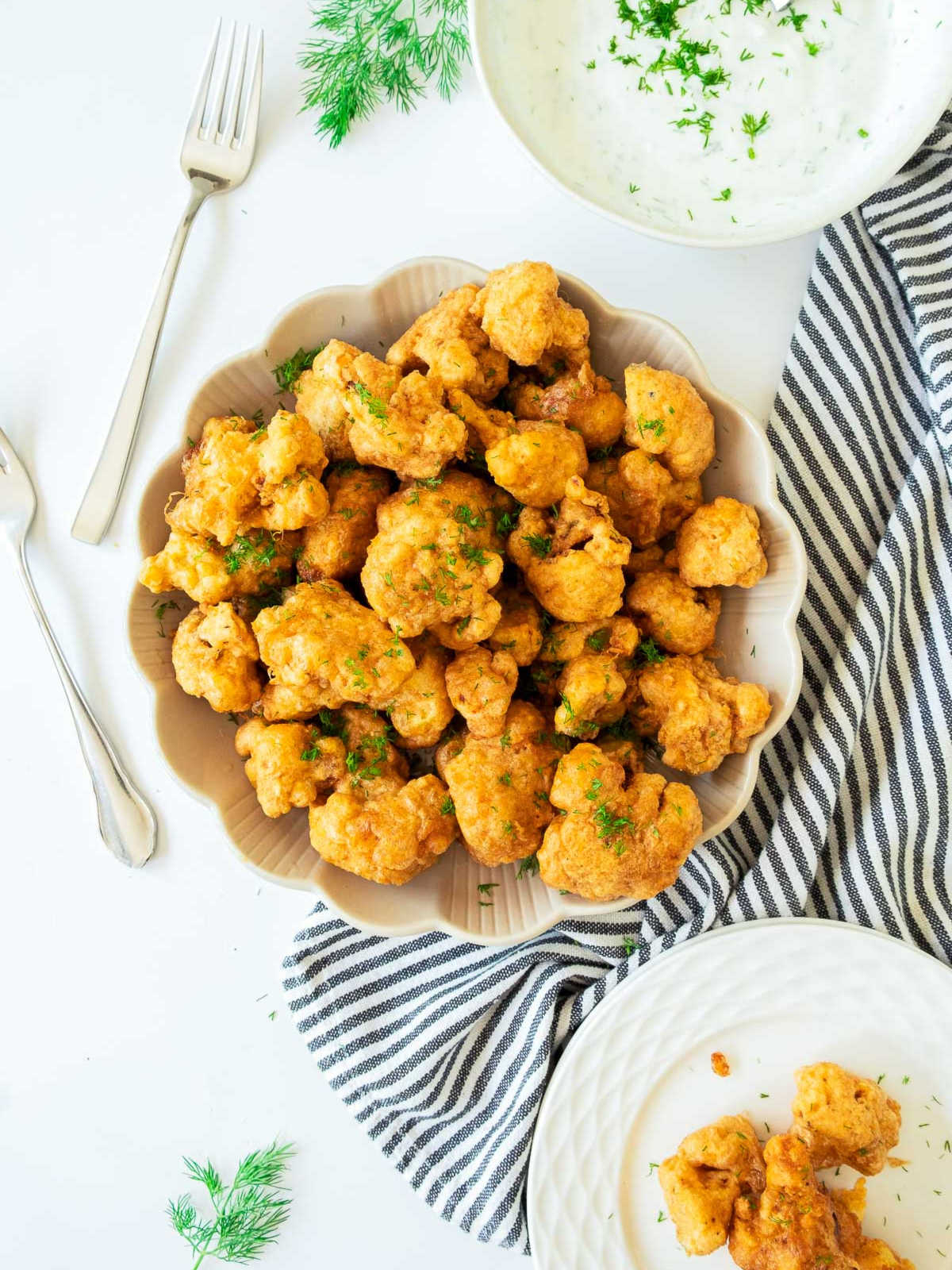 Overhead view of a generous serving of crispy, battered, and fried cauliflower bites (or wings) garnished with parsley, served in a scalloped beige bowl. A bowl of creamy white dipping sauce, forks, and a striped towel are arranged around the main dish.