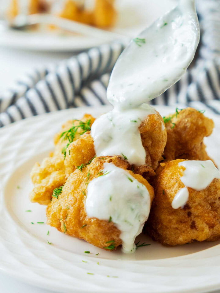 Close-up shot of crispy, golden-brown cauliflower bites being drizzled with a spoonful of creamy white dipping sauce, likely ranch or a dill yogurt sauce, on a white plate.