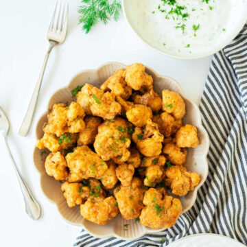 Overhead view of a generous serving of crispy, battered, and fried cauliflower bites (or wings) garnished with parsley, served in a scalloped beige bowl. A bowl of creamy white dipping sauce, forks, and a striped towel are arranged around the main dish.