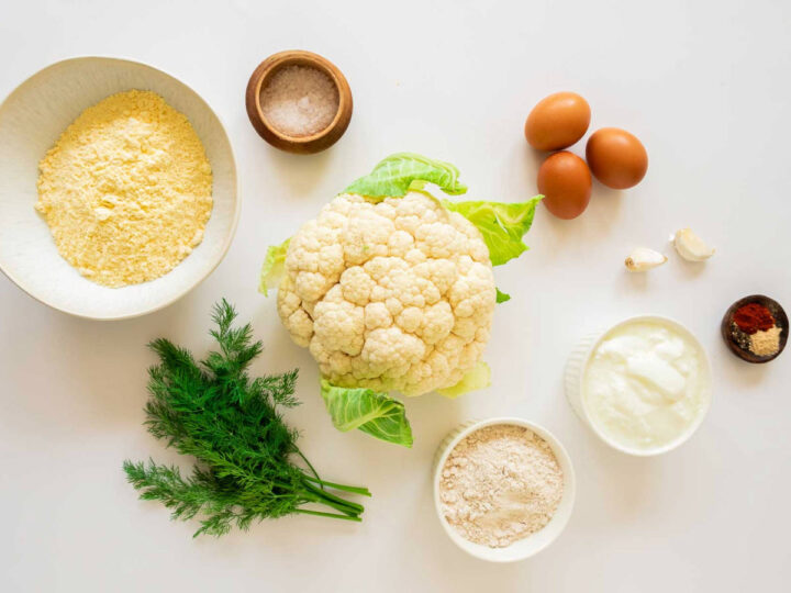 The image is an overhead flat lay showcasing the ingredients required to make crispy cauliflower bites: a whole head of cauliflower sits centrally, surrounded by bowls containing yellow flour (cornmeal/chickpea flour), a brown flour mixture, yogurt or sour cream, and a blend of spices (red pepper and light powder); accompanying these are three whole brown eggs, two cloves of garlic, a small pot of salt, and a bunch of fresh dill for seasoning and garnish.