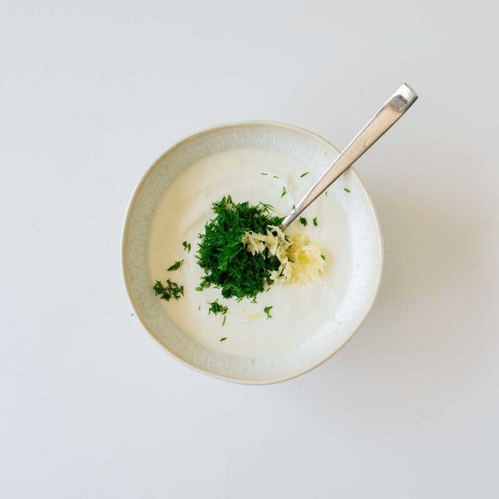 An overhead close-up shows a white bowl containing a creamy base (likely yogurt or sour cream) with a spoonful of finely chopped fresh dill and minced garlic placed in the center, ready to be mixed for a dipping sauce.